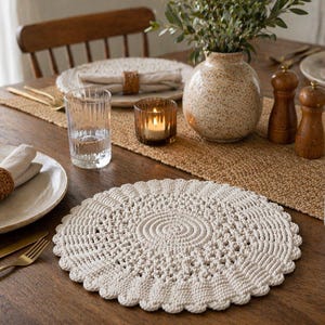 May include: A dining table set with a cream crocheted placemat. The table is set with plates, napkins, and gold cutlery. A vase with greenery, a candle, and salt and pepper shakers are also on the table.