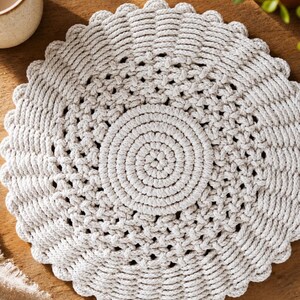 May include: A round, off-white coaster or placemat crafted from thick, braided cotton cord. The design features a solid, tightly coiled centre surrounded by an intricate, open-weave lattice pattern, finished with a scalloped, rounded edge.