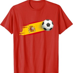 May include: A bright red short-sleeved t-shirt featuring a graphic of a soccer ball positioned next to a horizontal brushstroke design of the Spanish flag, which includes the national coat of arms centered on the yellow stripe.