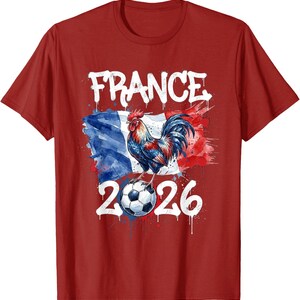 Op de afbeelding: Rood T-shirt met een aquarelontwerp. Het woord "FRANCE" staat in het wit boven een haan die op een voetbal staat. De Franse vlag staat op de achtergrond en het nummer "2026" staat eronder.