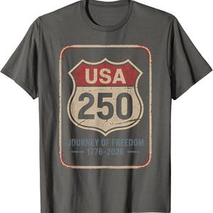 以下が含まれることがあります： グレーのTシャツ。前面には、赤とクリーム色の「USA 250」と書かれた道路標識のグラフィックがプリントされています。「Journey of Freedom 1776-2026」の文字が標識の下にあります。