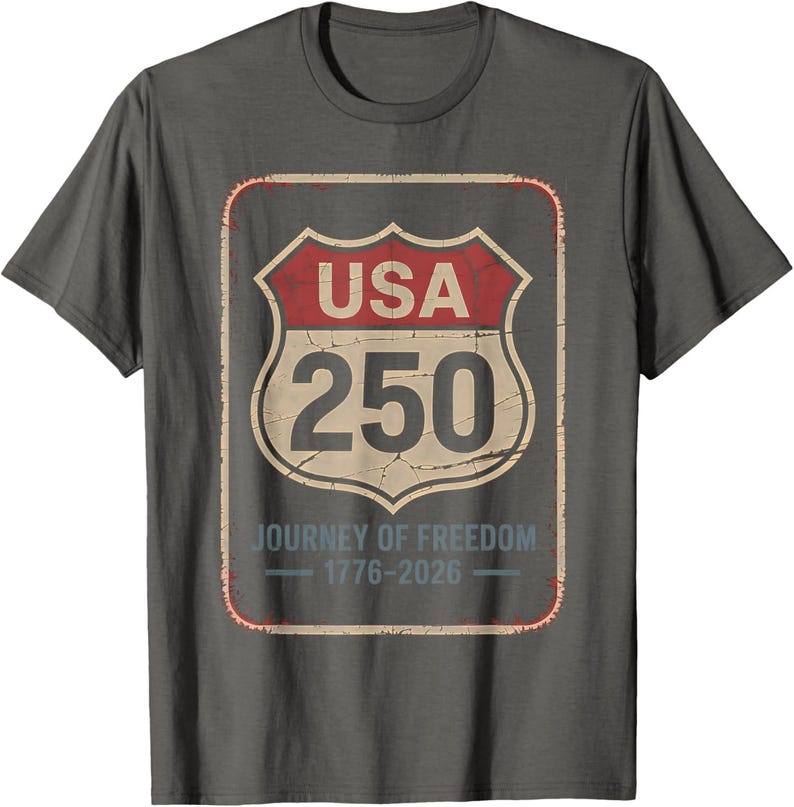 以下が含まれることがあります： グレーのTシャツ。前面には、赤とクリーム色の「USA 250」と書かれた道路標識のグラフィックがプリントされています。「Journey of Freedom 1776-2026」の文字が標識の下にあります。