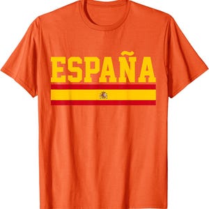 May include: An orange short-sleeved t-shirt featuring the word ESPAÑA in large yellow block letters. Below the text is a horizontal stripe design in red and yellow, mimicking the Spanish flag, with a small coat of arms centered in the middle.