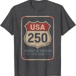 以下が含まれることがあります： 濃いグレーのTシャツ。USAの道路標識を模したかすれたグラフィックで、「USA 250」と「Journey of Freedom 1776-2026」の文字が入っています。デザインは赤、ベージュ、黒です。