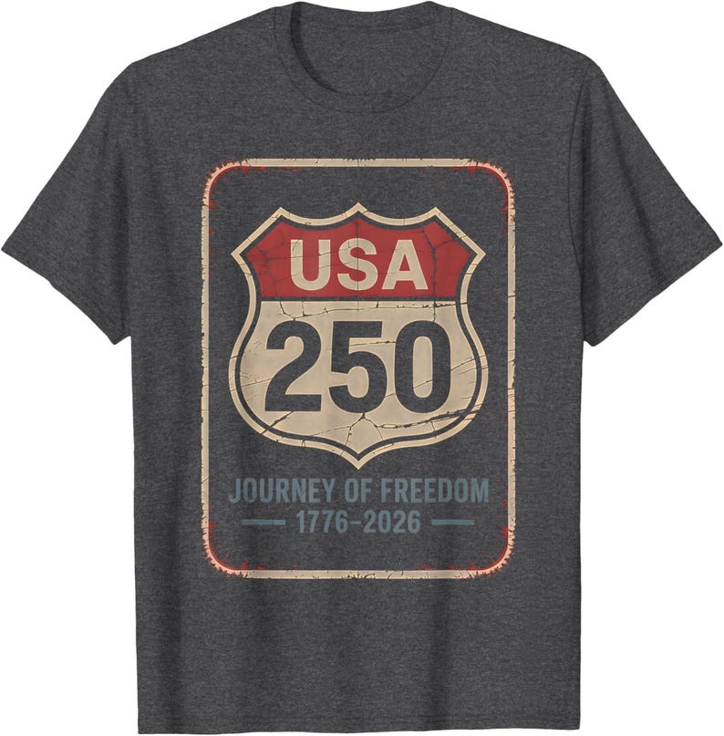 以下が含まれることがあります： 濃いグレーのTシャツ。USAの道路標識を模したかすれたグラフィックで、「USA 250」と「Journey of Freedom 1776-2026」の文字が入っています。デザインは赤、ベージュ、黒です。