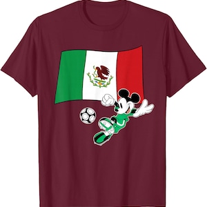 May include: Burgundy t-shirt featuring a graphic of Mickey Mouse in a green soccer uniform kicking a soccer ball. Behind Mickey is the Mexican flag. The design is centered on the front of the shirt.
