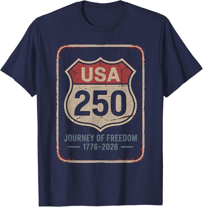 以下が含まれることがあります： ネイビーブルーのTシャツ。USA 250と「Journey of Freedom 1776-2026」と書かれた、使い古された道路標識のグラフィック。デザインは赤、ベージュ、白です。