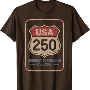 以下が含まれることがあります： 茶色のTシャツ。アメリカの道路標識のグラフィックがプリントされています。赤と白の標識には「USA 250」と「Journey of Freedom 1776-2026」と書かれています。クルーネックと半袖のデザインです。