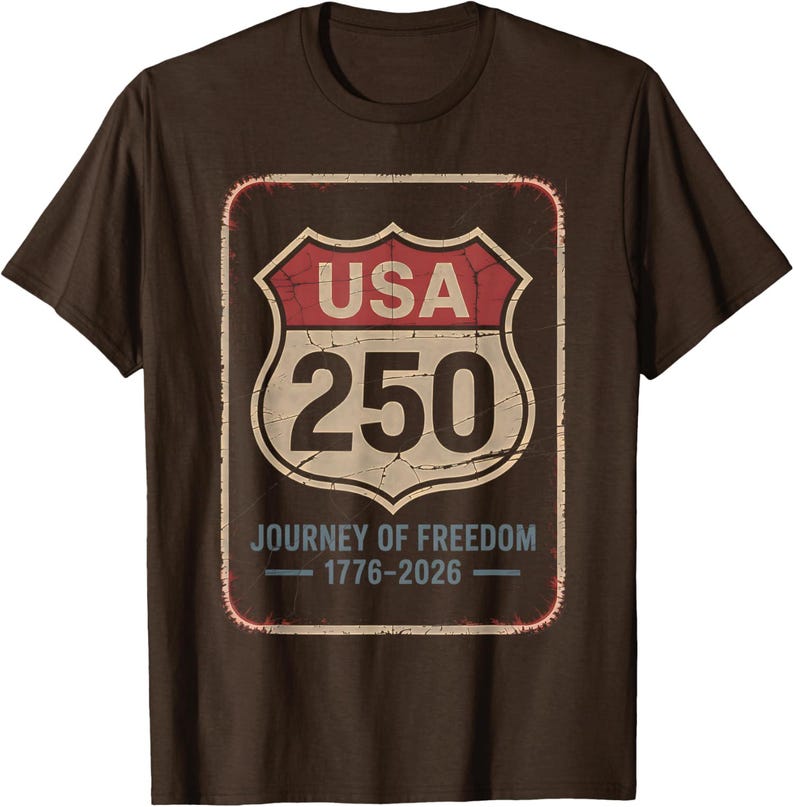以下が含まれることがあります： 茶色のTシャツ。アメリカの道路標識のグラフィックがプリントされています。赤と白の標識には「USA 250」と「Journey of Freedom 1776-2026」と書かれています。クルーネックと半袖のデザインです。