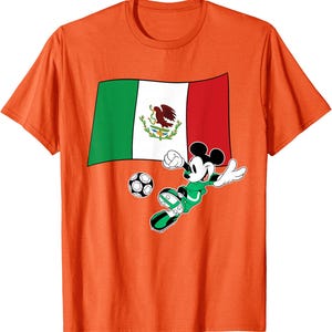 May include: Orange t-shirt featuring a graphic of Mickey Mouse playing soccer in a green uniform. The design includes the Mexican flag and a soccer ball. The shirt is made of a soft material.