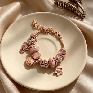 May include: Rose gold charm bracelet with pink and clear accents, featuring heart, flower, and gem-shaped charms. A matching rose gold hair clip sits nearby. The bracelet is displayed on a cream-colored plate.