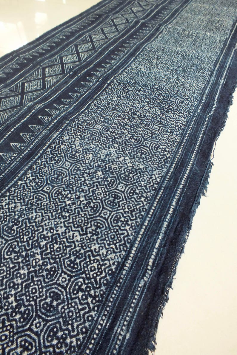 May include: A long, narrow piece of indigo-dyed fabric with a white geometric pattern. The fabric has a frayed edge and is folded in half.
