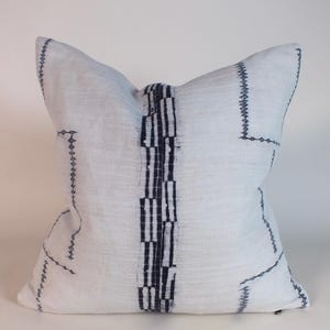 May include: White decorative pillow with a textured surface and dark blue stitched geometric patterns. The pillow features a central vertical stripe of alternating dark blue and white stripes. The pillow is square and has a soft, inviting appearance.