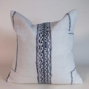 May include: White square pillow with a central vertical band and border stitching in dark blue. The pillow has a textured appearance and a unique shape, adding a decorative touch to any room. The pillow's design features geometric patterns.