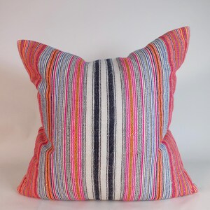 May include: A square pillow with a striped pattern in shades of red, pink, orange, blue, and white. The pillow has a textured appearance and is likely made of fabric. The pillow is a decorative item for a bed or sofa.