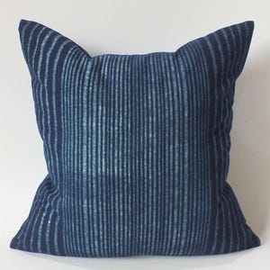 May include: A square throw pillow featuring a deep navy blue fabric with a vertical striped pattern. The stripes consist of varying widths of lighter blue and white, creating a textured, hand-dyed appearance across the entire surface of the pillow.