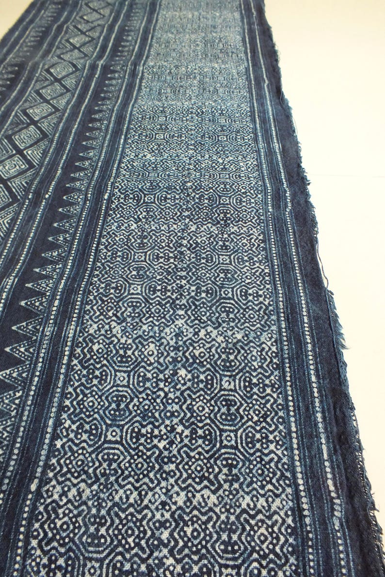 May include: A close-up of a blue and white indigo fabric with a geometric pattern. The fabric has a frayed edge and is woven with a fine texture.