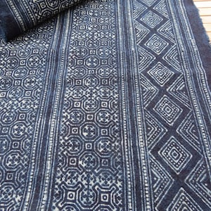 May include: Indigo blue fabric with geometric patterns. The fabric features repeating designs of diamonds, circles, and vertical stripes. The patterns are in white against a dark blue background. The fabric is partially rolled up, showing the texture and design.