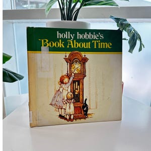 May include: A vintage children's book titled "Holly Hobbie's Book About Time." The cover features an illustration of two young girls and a black cat interacting with a grandfather clock. The book has a green and yellow banner at the top.