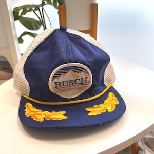 May include: A blue and white trucker cap featuring a circular patch with a mountain graphic and the text BUSCH. The cap includes a yellow braided cord across the base of the crown and two yellow embroidered leaf designs on the flat peak.