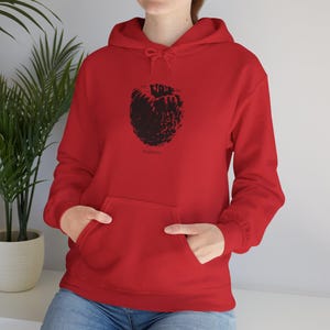 May include: A red hoodie featuring a black raspberry design. The word "raspberry" is printed beneath the fruit. The sweatshirt has a front pocket and a drawstring hood. The model is wearing blue jeans.