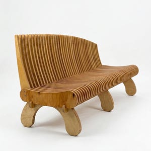 Puede incluir: Banco de madera con respaldo y asiento curvos, construido con listones de madera verticales y paralelos. El banco tiene cuatro patas curvas y es de madera marrón claro. El diseño es moderno y minimalista.