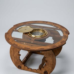 Può includere: Un tavolo rotondo in legno con piano in vetro dotato di un inserto centrale circolare con bordo in ottone e un coperchio in ottone abbinato. La base è composta da pannelli in legno a incastro con intagli e intarsi decorativi.