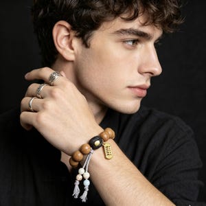 May include: A wooden bead bracelet with a black accent bead and a gold-coloured charm. The bracelet features a grey tassel and smaller beads in white and blue. The person is also wearing silver rings.