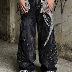 Op de afbeelding: Zwarte en grijze distressed denim jeans met een wijde pijp, gerafelde randen en contrasterende lichtgrijze panelen. De broek is voorzien van een zwarte riem met een zilverkleurige metalen ketting, vetersluiting aan de zijkanten en meerdere zakken.