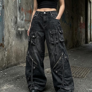 Puede incluir: Pantalones cargo de mezclilla negros con múltiples bolsillos y cremalleras. Los pantalones tienen un diseño de pierna ancha y se usan con una camiseta sin mangas negra y zapatos negros. Los pantalones tienen un aspecto descolorido y desgastado.