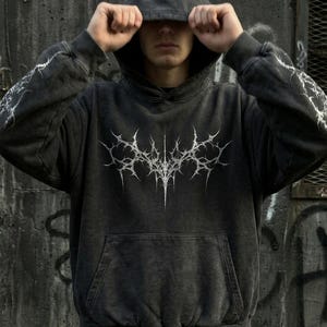May include: Dark gray hoodie with a white, spiky graphic on the front and lightning bolt designs on the sleeves. The hoodie has a drawstring hood and a front pocket. The garment is made of a soft, textured material.