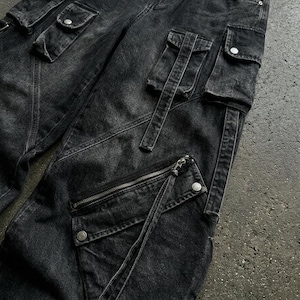 A close-up detail shot of the black denim fabric, flap pockets, and diagonal zipper of the cargo pants on a textured surface.
