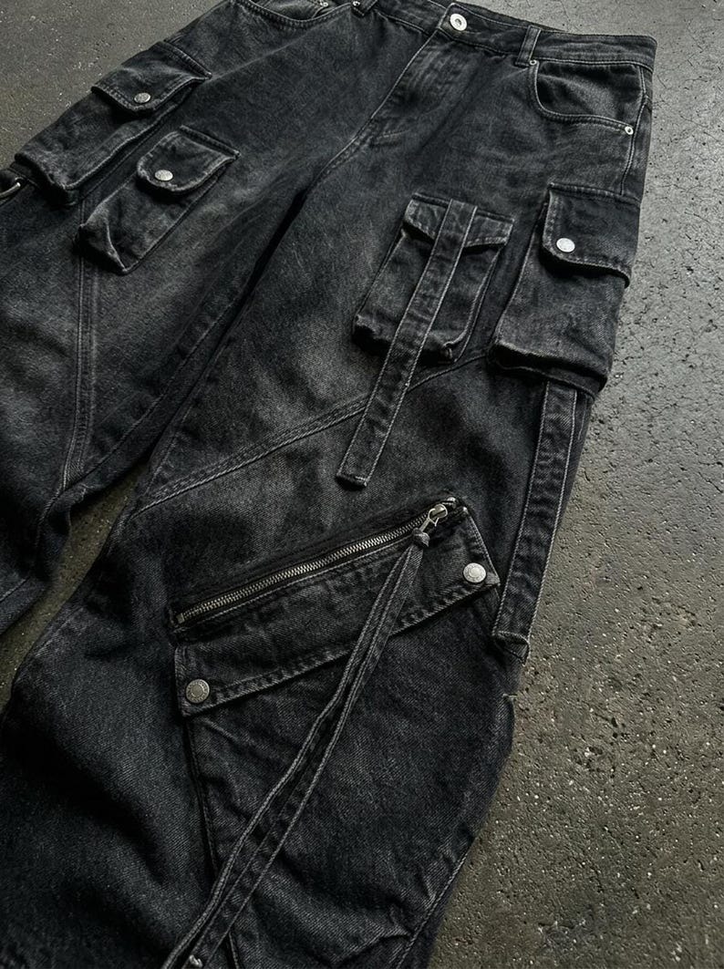 A close-up detail shot of the black denim fabric, flap pockets, and diagonal zipper of the cargo pants on a textured surface.