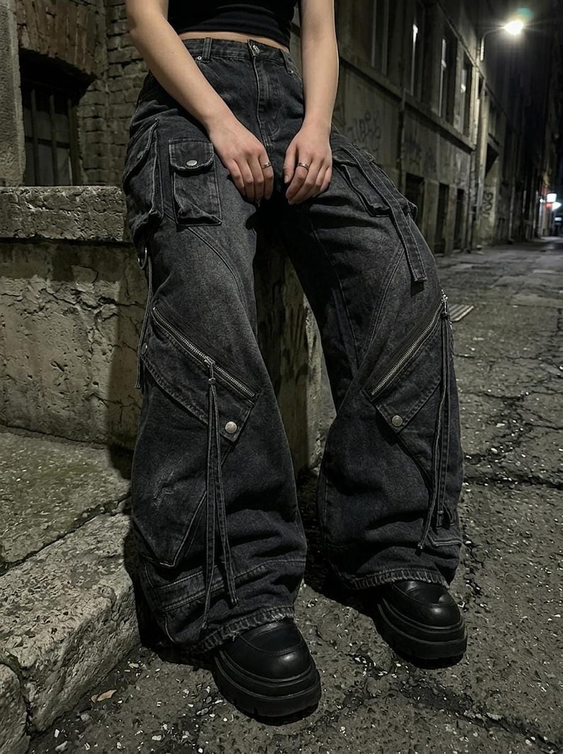 A person sitting on a concrete ledge at night wearing black utility cargo denim pants with prominent pockets and thick-soled shoes.