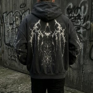 May include: Dark gray hoodie with a white, intricate graphic design on the back. The hoodie has a hood and a relaxed fit. The design appears to be a stylized, abstract pattern. The person is wearing black pants.