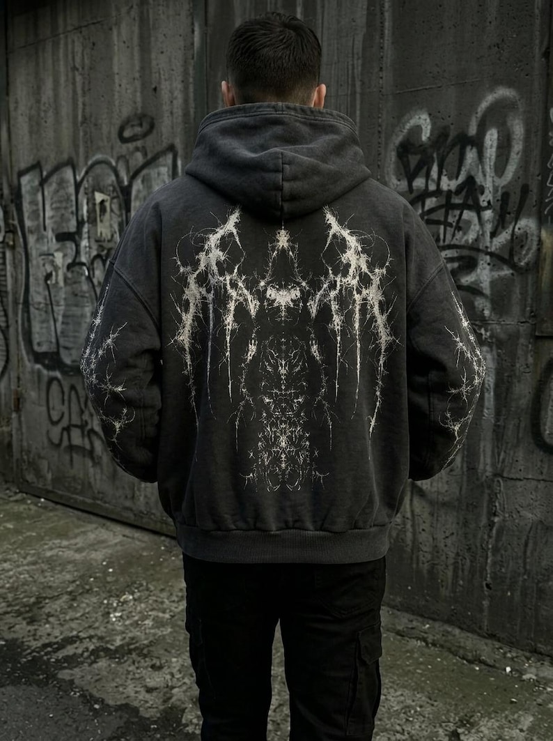 May include: Dark gray hoodie with a white, intricate graphic design on the back. The hoodie has a hood and a relaxed fit. The design appears to be a stylized, abstract pattern. The person is wearing black pants.