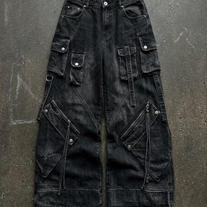 A flat lay image of black wide-leg denim cargo pants featuring multiple flap pockets, zippers, and straps laid out on a dark concrete surface.