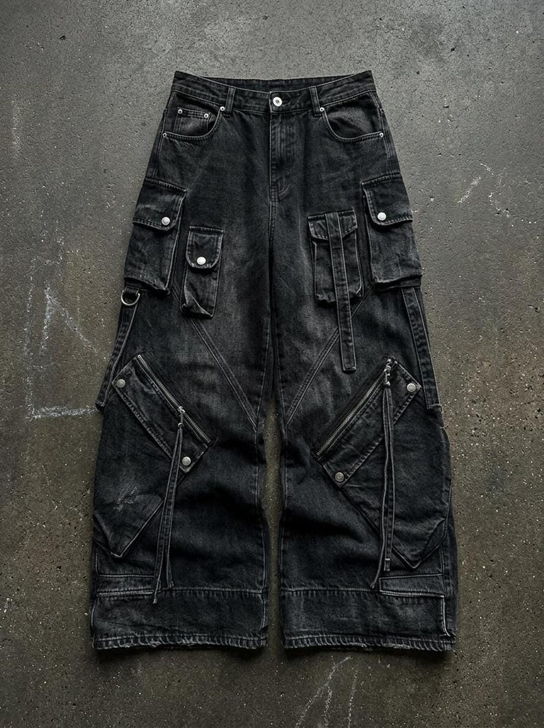 A flat lay image of black wide-leg denim cargo pants featuring multiple flap pockets, zippers, and straps laid out on a dark concrete surface.