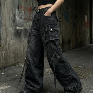 A side-angle view of a person wearing black distressed cargo jeans with prominent side pockets and straps in an urban alley.