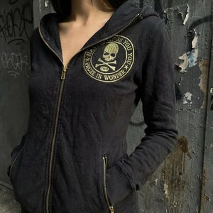 Peut inclure: Sweat à capuche noir zippé avec un emblème doré de tête de mort et d'os croisés. L'emblème est entouré des mots "FROZE IN WONDER" et "YOU YOU". Le sweat à capuche a une capuche, une fermeture éclair et deux poches avant.