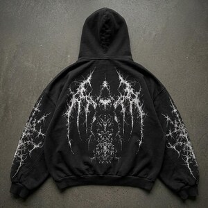 May include: Black hoodie with a white, intricate, spiky design on the back and sleeves. The design has a gothic or abstract aesthetic. The hoodie has a hood and ribbed cuffs and hem.