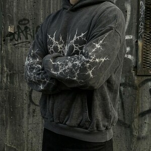 May include: Dark gray hoodie with a white lightning bolt design on the chest and sleeves. The hoodie has a front pocket and a drawstring hood. The garment is made of a soft, washed fabric.
