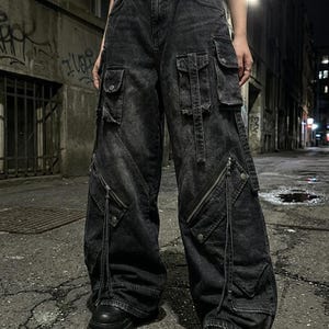 A close-up of the front of a person wearing black baggy cargo pants with diagonal zippered pockets, standing on a paved street at night.