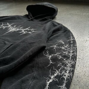 May include: Dark gray hoodie with a hood and a front pocket. The hoodie features a white, spiky graphic design on the chest and down the right sleeve. The fabric has a slightly faded appearance.