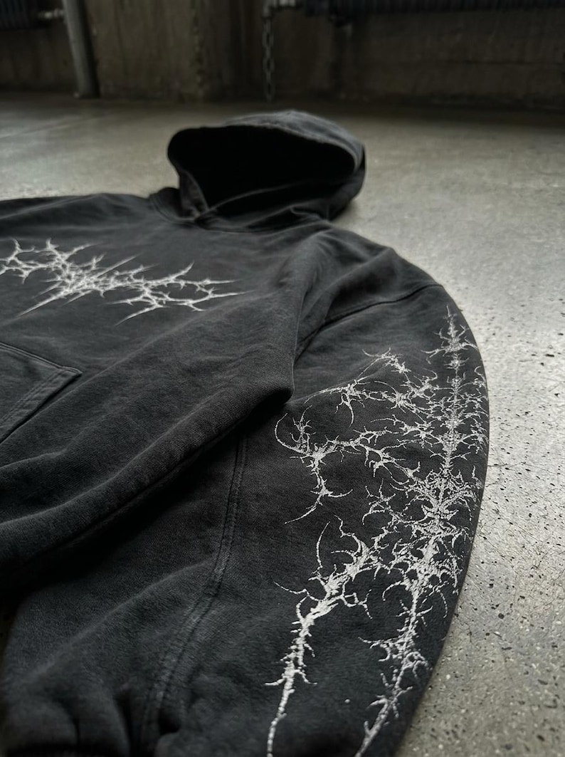 May include: Dark gray hoodie with a hood and a front pocket. The hoodie features a white, spiky graphic design on the chest and down the right sleeve. The fabric has a slightly faded appearance.