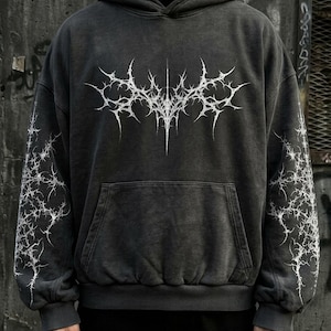 May include: Dark gray hoodie with a white, spiky graphic on the front and sleeves. The hoodie has a kangaroo pocket and a drawstring hood. The fabric has a washed, vintage look. The design is a gothic or edgy style.