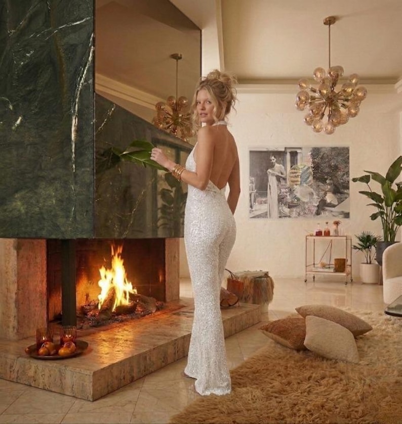May include: A floor-length white sequined jumpsuit featuring a halter neckline and an open back design. The garment is covered in shimmering sequins that reflect light, creating a textured appearance throughout the fitted bodice and flared legs.