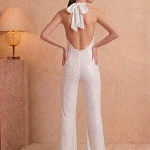 K&ouml;nnte beinhalten: Ein wei&szlig;er Pailletten-Jumpsuit mit r&uuml;ckenfreiem Design und einer gro&szlig;en Stoffschleife im Nacken. Das Kleidungsst&uuml;ck hat einen Neckholder-Ausschnitt und lange, ausgestellte Hosenbeine. Ein wei&szlig;er floraler Kopfschmuck wird im Haar getragen.