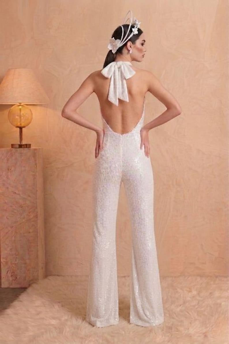 K&ouml;nnte beinhalten: Ein wei&szlig;er Pailletten-Jumpsuit mit r&uuml;ckenfreiem Design und einer gro&szlig;en Stoffschleife im Nacken. Das Kleidungsst&uuml;ck hat einen Neckholder-Ausschnitt und lange, ausgestellte Hosenbeine. Ein wei&szlig;er floraler Kopfschmuck wird im Haar getragen.