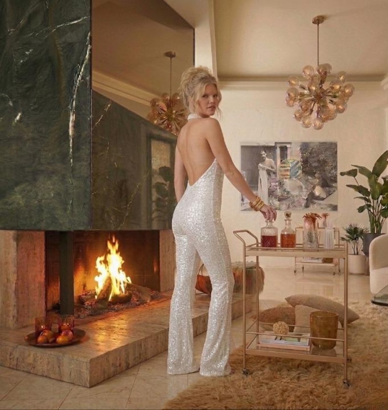 May include: A white sequined jumpsuit featuring a halter neckline and an open back design. The garment has a fitted bodice that transitions into flared pant legs, covered entirely in shimmering white sequins that reflect the light.